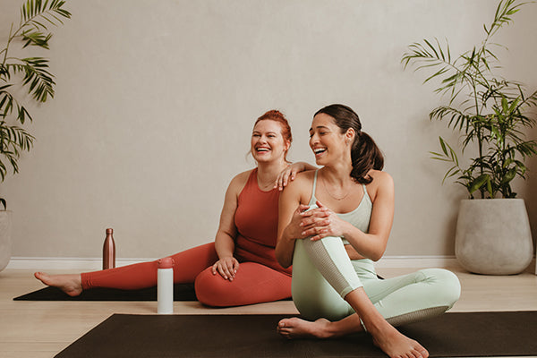 Two women sitting on yoga mats, smiling, energy and workout concept